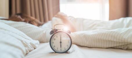 alarma reloj con asiático mujer en cama mientras durmiendo, joven adulto hembra despertar arriba tarde en el temprano Mañana. relajante , somnoliento, diario rutina y tener un bonito día conceptos foto