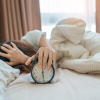 alarm clock and asian woman hand stop time in bed while sleeping, young adult female wake up late in the early morning. Relaxing , sleepy, daily routine and have a nice day concepts photo
