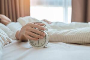 alarm clock and asian woman hand stop time in bed while sleeping, young adult female wake up late in the early morning. Relaxing , sleepy, daily routine and have a nice day concepts photo