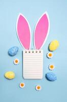 Easter concept. Notepad with place for text with rabbit ears and easter eggs on blue background. Flat lay, top view, copy space. photo