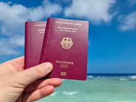 A hand holds two German passports in front of a soft travel background in the Maldives with palm trees and beach. photo