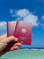 A hand holds two German passports in front of a soft travel background in the Maldives with palm trees and beach. photo