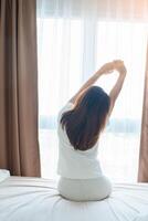 contento mujer extensión en cama después despertar arriba, joven adulto hembra creciente brazos y mirando a ventana en el temprano Mañana. Fresco relajarse y tener un bonito día conceptos foto