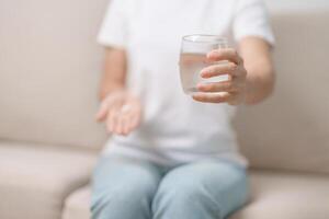 mujer mano participación medicina analgésico píldora y agua vaso en el sofá a hogar, tomando para dolores de cabeza, estómago dolor, Diarrea dolor desde comida envenenamiento, endometriosis, histerectomía y menstrual foto