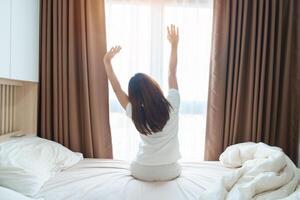 happy woman stretching on bed after wake up, young adult female rising arms and looking to window in the early morning. fresh relax and have a nice day concepts photo