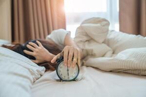 alarma reloj y asiático mujer mano detener hora en cama mientras durmiendo, joven adulto hembra despertar arriba tarde en el temprano Mañana. relajante , somnoliento, diario rutina y tener un bonito día conceptos foto