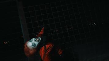a man in a red shirt and clown makeup dances in front of an iron fence while posing very creepily at night. video