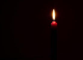 Single burning candle flame or light glowing on a small red candle on black or dark background on table in church for Christmas, funeral or memorial service with copy space photo