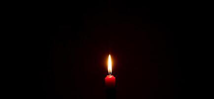 A single burning candle flame or light glowing on a red candle on black or dark background on table in church for Christmas, funeral or memorial service with copy space photo