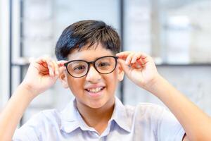 Smiling Indian-thai boy choosing glasses in optics store, Portrait of Mixed race ethnicity kid wearing glasses at the optical store photo
