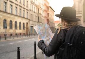 young girl is looking at a city map on the street on a journey. woman has blond hair wearing a leather jacket, hat and backpack.new travel concept in new realities. High quality photo