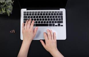 Black office desk with laptop. Hands typing on the keyboard. flower stands nearby. Top view with copy space for text input. High quality photo