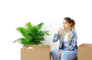 Tired young woman sits among unpacked cardboard boxes with a glass of water.Fern flower stands nearby.moving to a new house. High quality photo