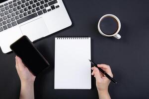 hand holds pen over notebook on black table near laptop.left hand holding a smartphone. cup of coffee nearby. glasses are on computer.Top view with copy space for text input. photo