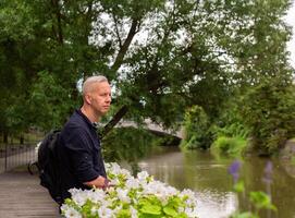 gray-haired man with a backpack leaning on a fence near the river and looking into the distance photo