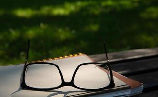 lentes mentira en parte superior de un pila de libros con un cuaderno en un banco en el parque en un soleado día en el sombra de arboles en verano. selectivo foco.alto calidad foto