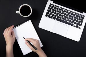 hand holds pen over notebook on black table near laptop. cup of coffee nearby. Top view with copy space for text input. photo