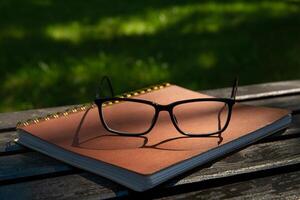 glasses lie on a leather notebook on a park bench on a sunny day in the shade of trees in summer. background blurred. selective focus photo