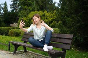 happy Caucasian woman sitting on a park bench on a sunny day, holding a phone in her hand, talking on video chat and waving her hand photo