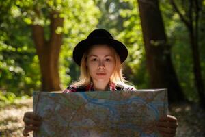 joven caucásico mujer en un sombrero mirando a un geográfico mapa en el sombra de arboles en un parque o bosque. alto calidad foto
