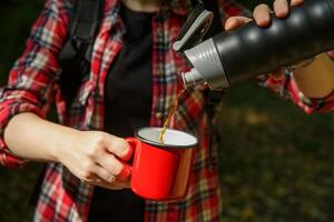 turista manos verter café desde un termo dentro un metal rojo copa.selectiva enfocar. antecedentes borroso. alto calidad foto