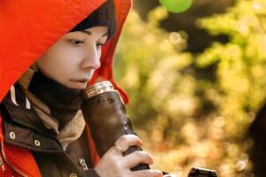 a young woman in a red jacket holds a thermos with a hot drink at her face and inhales the aroma. High quality photo