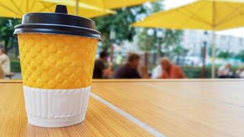 uno amarillo papel vaso de té o café - para llevar comida en un mesa en un café tienda. nadie. un desechable vaso soportes en contra el fondo de un verano café con irreconocible gente. foto