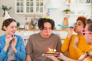 Make a wish. Man wearing party cap blowing out burning candles on birthday cake. Happy Birthday party. Group of friends wishes guy happy birthday. People celebrating birthday with party at home photo