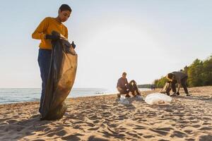 tierra día. voluntarios activistas recoge basura limpieza de playa costero zona. mujer y mans pone el plastico basura en basura bolso en Oceano costa. ambiental conservación costero zona limpieza foto