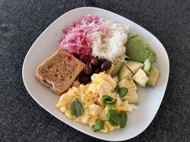 Homemade breakfast with avocado, scrambled eggs, olives, kohlrabi, reddish and a slice of bread served on a white plate photo