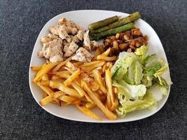 hecho en casa A la parrilla pollo con francés papas fritas, espárragos, A la parrilla Cortado berenjena y verde ensalada, servido en un blanco plato foto