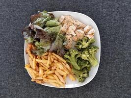 hecho en casa A la parrilla pollo con francés papas fritas, brócoli y verde ensalada, servido en un blanco plato foto