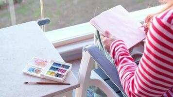 dibujar en bloc de dibujo. espalda ver de un mujer dibujar en bloc de dibujo sentado en el balcón video