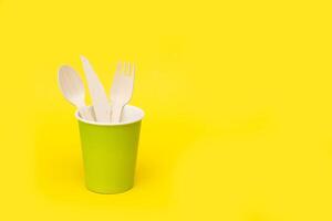 disposable wooden spoon, fork and knife in a green paper cup on a yellow background with copy space, recycled. photo