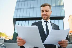 Cheerful young middle-eastern man entrepreneur getting ready before presentation, sitting on the street by office building with backpack, holding papers and drinking coffee, copy space photo