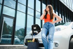 Young woman is standing near the electric car and looks at the smart phone. The rental car is charging at the charging station for electric vehicles. Car sharing. photo