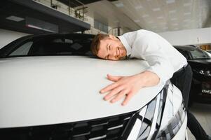 Happy handsome bearded man buying a car in dealership, guy hugging hood of new car photo