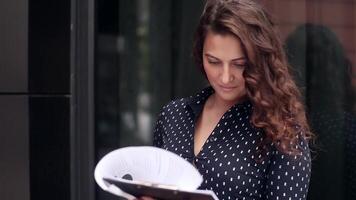 Close-up. Young attractive business woman in dark shirt and beautiful hair looks office documents near office building. Business project. Business plan video