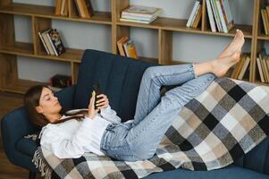 cerca arriba de un relajado niña utilizando un inteligente teléfono acostado en un sofá en el vivo habitación a hogar. foto
