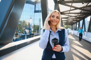 hermosa mujer yendo a trabajo con café caminando cerca oficina edificio. retrato de exitoso negocio mujer participación taza de caliente beber. foto