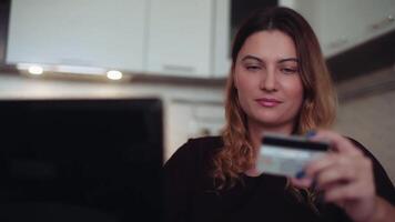 mooi jong vrouw met lang donker haar- online bank gebruik makend van een mobiel computer Aan de internet met een credit kaart Bij huis Bij de tafel hebben een mooi zo humeur en lachend. hd. huis stijl. video