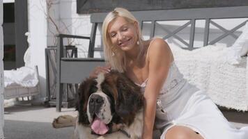 Young blonde girl hugs a big dog and smiles while sitting on a white porch near the house. Portrait video