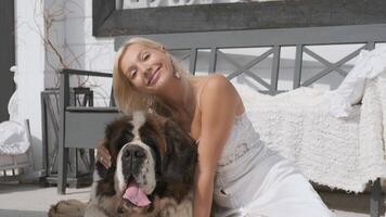 ung blond flicka kramar en stor hund och ler medan Sammanträde på en vit veranda på Hem video
