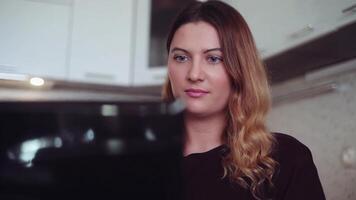 Close-up. Beautiful young business woman with long hair prints on a mobile computer in black at home sitting at a table in a good mood. video