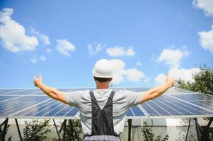 Smiling worker with solar station, raising his hands, showing thumbs up on a background of photovoltaic panels near the house. Man in orange uniform. Science solar energy. Renewable energy photo