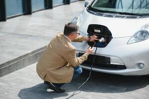 Hansome bearded man sitting near his new modern electric car and holding plug of the charger, while car is charging at the charging station photo