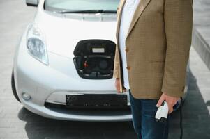 handsome man holding charging cable at electric charging station point standing near his new car. photo