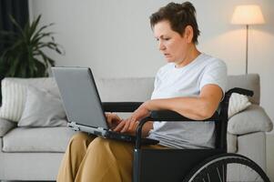 Middle age grey-haired disabled woman having video call sitting on wheelchair at home. photo