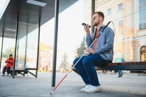 joven ciego hombre con teléfono inteligente sentado en banco en parque en ciudad, vocación. foto