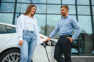 Happy young adult man and smiling woman charging electric car. photo